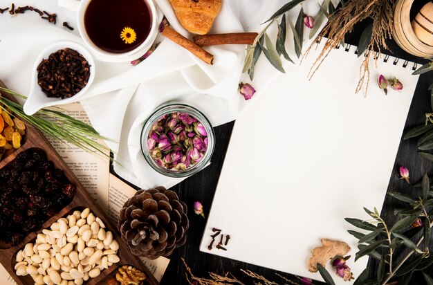 Bovenaanzicht van een schetsboek met theeroosknoppen in een glazen pot, dennenappels, gemengde noten en boombrunches met bladeren en een kopje thee op zwart hout