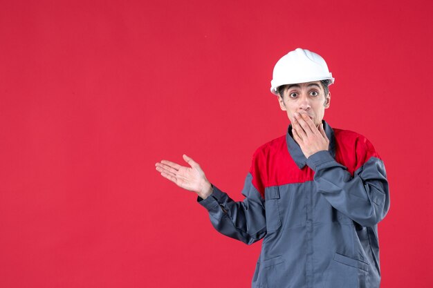 Bovenaanzicht van een verraste jonge werknemer in uniform met een helm die naar de rechterkant wijst op een geïsoleerde rode muur
