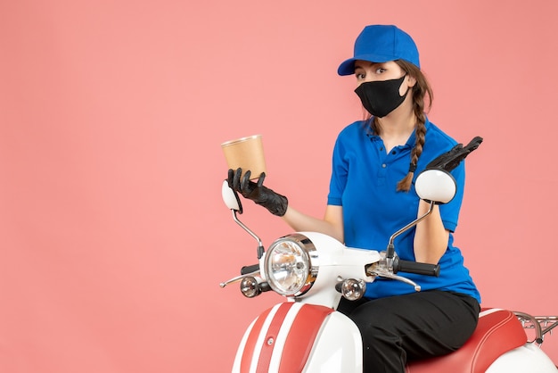 Bovenaanzicht van een verraste vrouwelijke koerier met een medisch masker en handschoenen die op een scooter zitten en bestellingen afleveren op pastel perzik