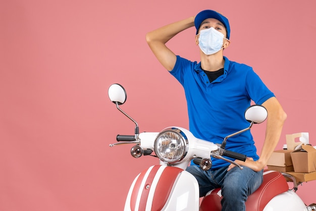 Bovenaanzicht van een verwarde koeriersman met een medisch masker met een hoed op een scooter op een pastelkleurige perzikachtergrond