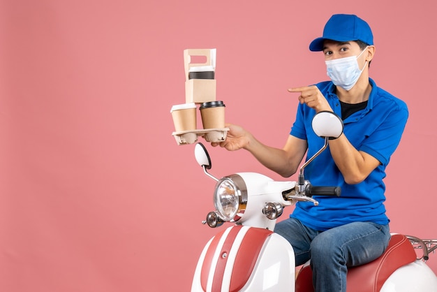 Bovenaanzicht van een zelfverzekerde mannelijke bezorger met een masker met een hoed op een scooter die bestellingen aflevert op een perzikachtergrond