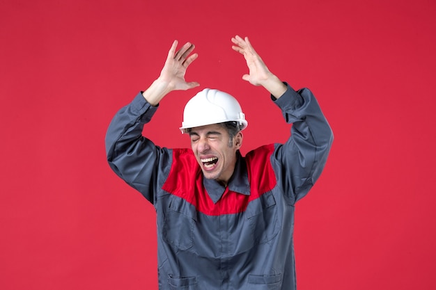 Bovenaanzicht van emotionele jonge bouwer in uniform met harde hoed en handen opstekend op geïsoleerde rode muur