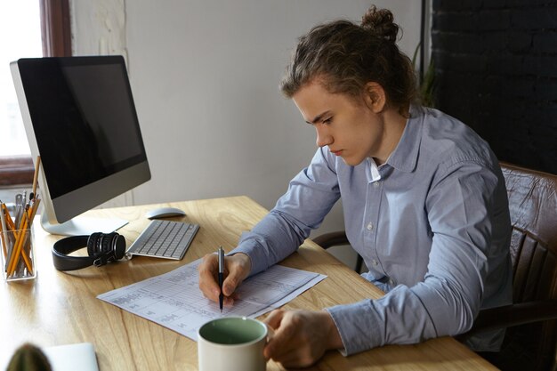 Bovenaanzicht van ernstige gerichte aantrekkelijke jonge mannelijke ingenieur met haar broodje zit op houten bureau voor draagbare computer met lege copyspace-scherm, tekeningen of specificaties controleren