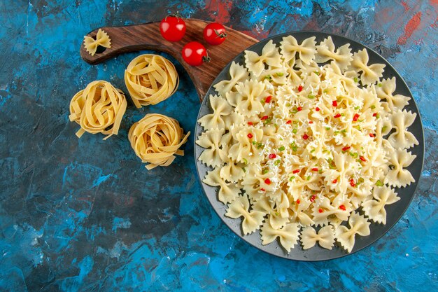 Bovenaanzicht van farfalle pasta's met groenten op een zwarte plaat naast tomaten op houten snijplank op blauwe achtergrond