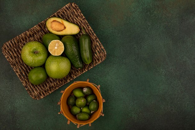 Bovenaanzicht van feijoas op een emmer met groene appels, avocado's, komkommer op een rieten dienblad op een groen oppervlak met kopie ruimte