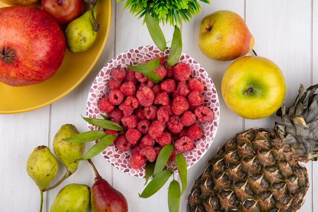 Bovenaanzicht van frambozen in kom met granaatappel ananas perzik en appel rond op een witte ondergrond