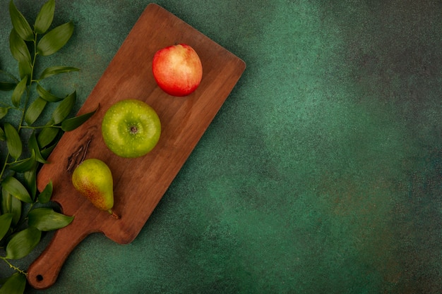 Bovenaanzicht van fruit als appel-perzik-peer op snijplank met bladeren op groene achtergrond met kopie ruimte