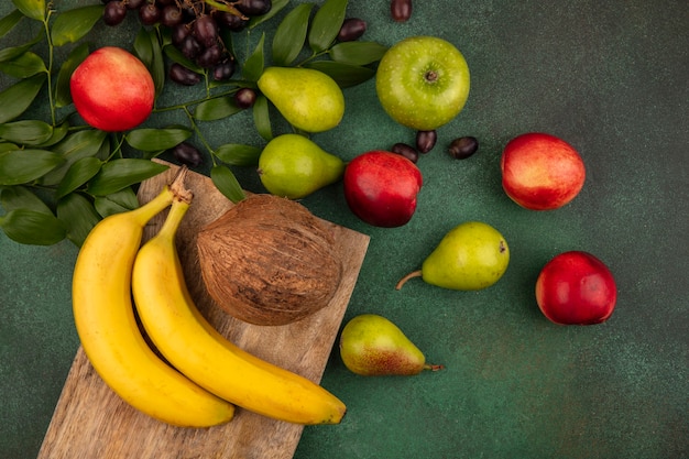 Bovenaanzicht van fruit als banaan kokosnoot op snijplank en druif perzik appel peer met bladeren op groene achtergrond