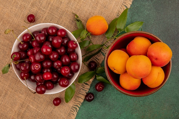 Bovenaanzicht van fruit als kersen en abrikozen in kommen met bladeren op zak op groene achtergrond