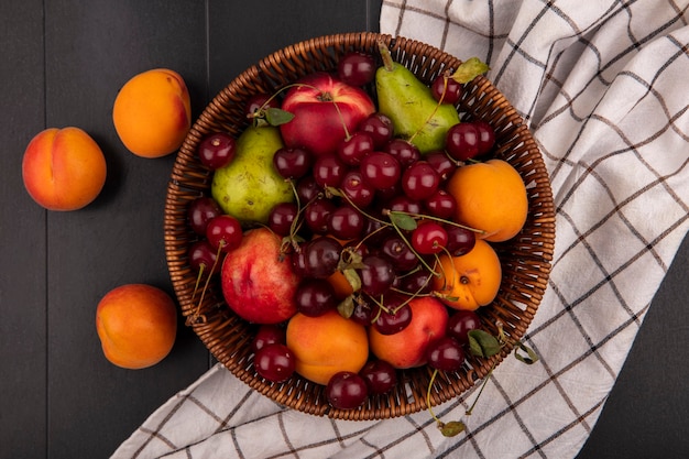 Bovenaanzicht van fruit als kersen perzik abrikoos peer in mand op geruite doek en op zwarte achtergrond