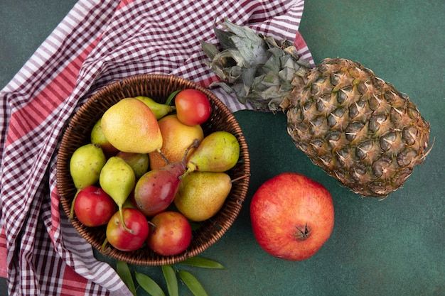 Bovenaanzicht van fruit als perzik appel pruim in mand op geruite doek met granaatappel en ananas op groene ondergrond