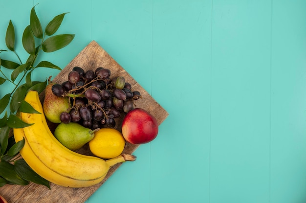 Bovenaanzicht van fruit als perzik peer citroen druif banaan met bladeren op snijplank op blauwe achtergrond met kopie ruimte