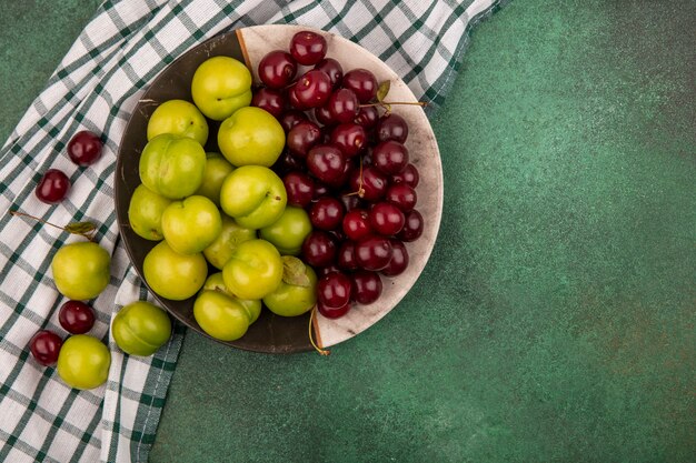 Bovenaanzicht van fruit als pruimen en kersen in plaat en op geruite doek op groene achtergrond met kopie ruimte