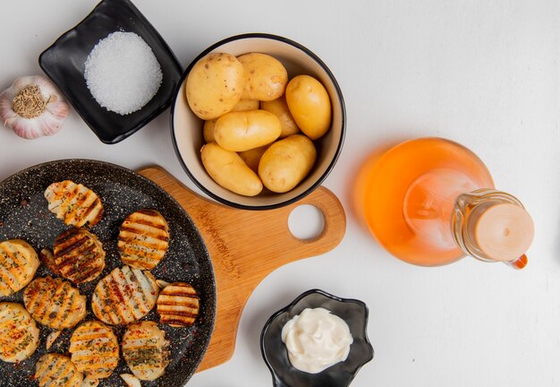 Bovenaanzicht van gebakken aardappelschijfjes in koekenpan op snijplank met ongekookte degenen in kom knoflook gesmolten boter mayonaise zout en zwarte peper op wit
