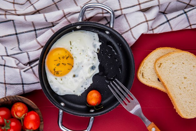 Bovenaanzicht van gebakken ei met tomaat en vork in pan op geruite doek en sneetjes brood kom tomaten op rood
