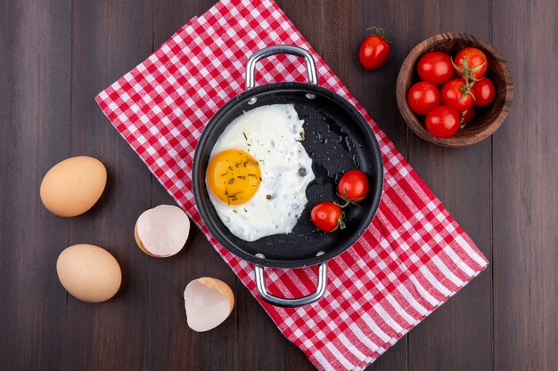 Bovenaanzicht van gebakken ei met tomaten in pan op geruite doek en eieren met shell en kom tomaat op houten oppervlak