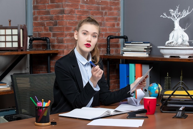 Bovenaanzicht van geconcentreerde serieuze jonge vrouw die aan een tafel zit en een document op kantoor vasthoudt