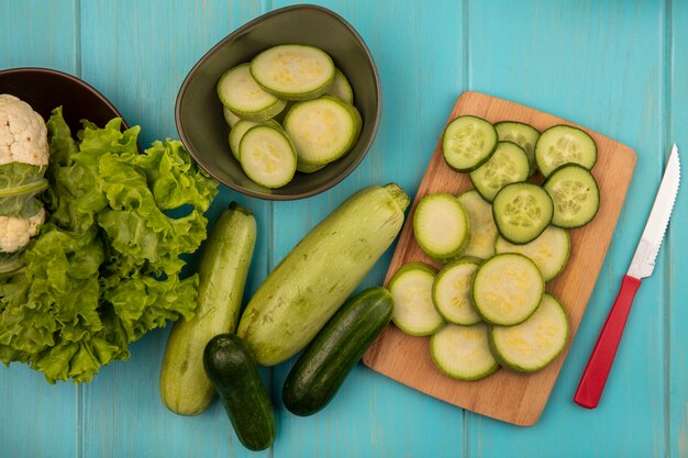 Bovenaanzicht van gehakte courgettes en komkommers op een houten keukenbord met mes met bloemkool en sla op een kom met hele komkommers en courgettes geïsoleerd op een blauwe houten ondergrond