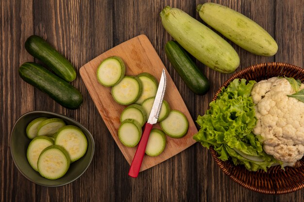 Bovenaanzicht van gehakte courgettes op een kom met gehakte courgettes op een houten keukenbord met mes met bloemkool en sla op een emmer met komkommer en courgettes geïsoleerd op een houten muur