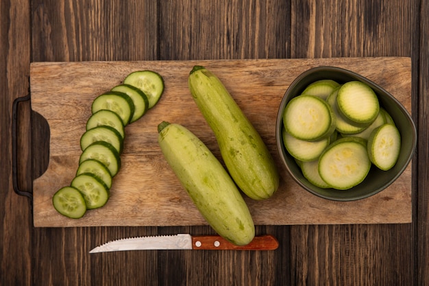 Bovenaanzicht van gehakte komkommers en courgettes geïsoleerd op een houten keukenplank met mes op een houten achtergrond