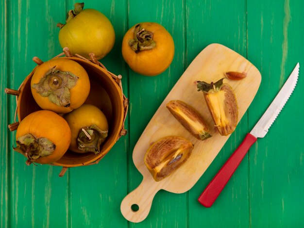 Bovenaanzicht van gehalveerde kaki fruit op een houten keukenplank met mes met kaki fruit op een emmer op een groene houten tafel