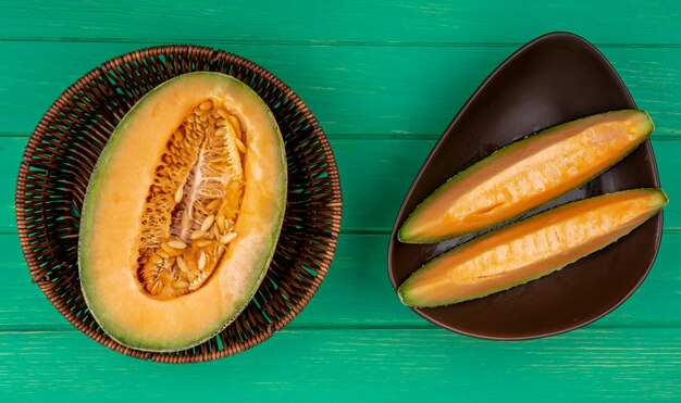 Bovenaanzicht van gehalveerde meloen meloen op een emmer met plakjes op een bruine kom op groene ondergrond