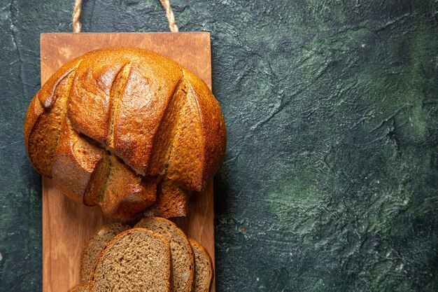 Bovenaanzicht van geheel en gesneden zwart brood op bruine houten snijplank aan de rechterkant op donkere kleuren achtergrond