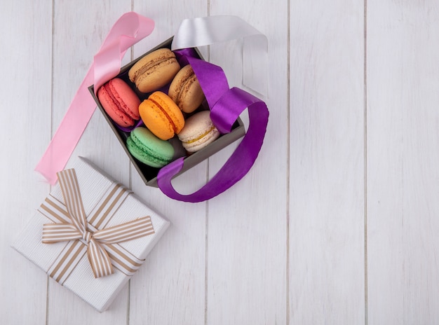 Bovenaanzicht van gekleurde macarons in een doos met gekleurde strikken en cadeaupapier op een wit oppervlak