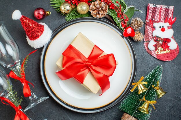 Bovenaanzicht van geschenkdoos op diner plaat kerstboom fir takken conifeer kegel kerstman hoed gevallen glazen bekers op zwarte achtergrond