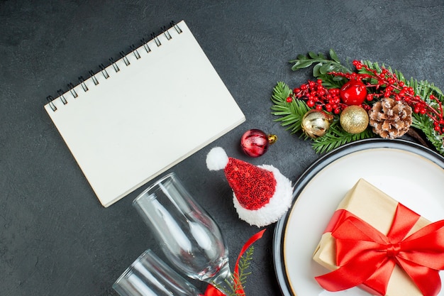 Bovenaanzicht van geschenkdoos op diner plaat kerstboom fir takken conifer kegel kerstman hoed gevallen glazen bekers notebook op zwarte achtergrond