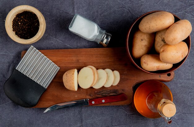 Bovenaanzicht van gesneden aardappel en mes met chips cutter op snijplank met andere in kom zout zwarte peper boter op grijze doek oppervlak