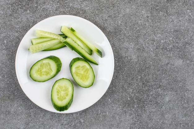 Bovenaanzicht van gesneden en gehakte plakjes komkommer op witte plaat over grijze tafel.