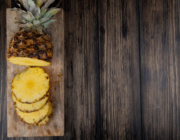 Bovenaanzicht van gesneden en gesneden ananas op snijplank aan de linkerkant en houten achtergrond met kopie ruimte