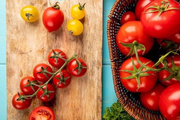 Bovenaanzicht van gesneden en hele tomaten op snijplank met andere in mand en koriander op blauw