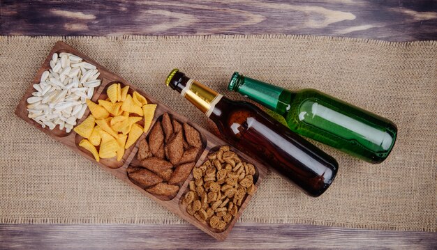 Gratis foto bovenaanzicht van gevarieerd bier snacks brood crackers chips en zonnebloempitten op een houten schotel met flessen bier op zak op rustiek