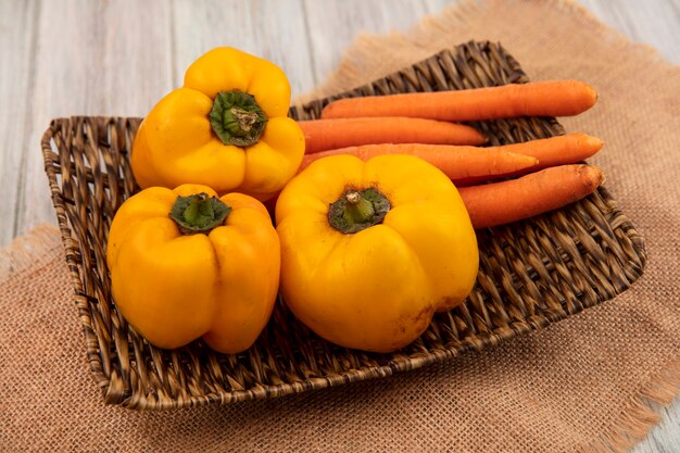 Bovenaanzicht van gezonde gele paprika met wortelen op een rieten dienblad op een zakdoek op een grijze houten ondergrond