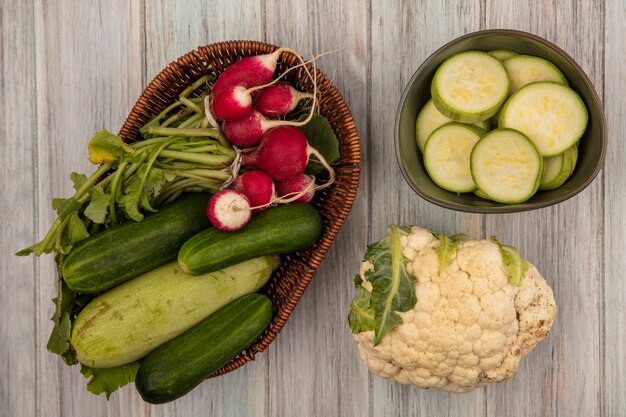 Bovenaanzicht van gezonde groenten zoals komkommers, courgettes en radijs op een emmer met bloemkool geïsoleerd op een grijze houten achtergrond