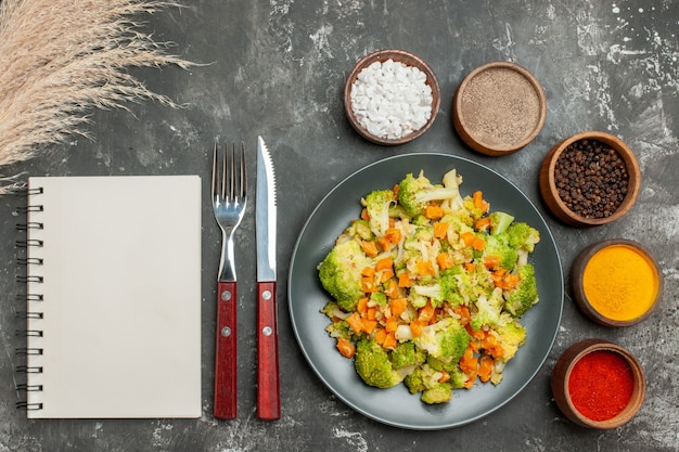 Bovenaanzicht van gezonde maaltijd met brocoli en wortelen op een zwarte plaat en kruiden op grijze achtergrond