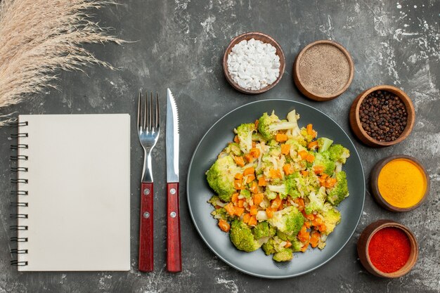 Bovenaanzicht van gezonde maaltijd met brocoli en wortelen op een zwarte plaat en kruiden op grijze achtergrond