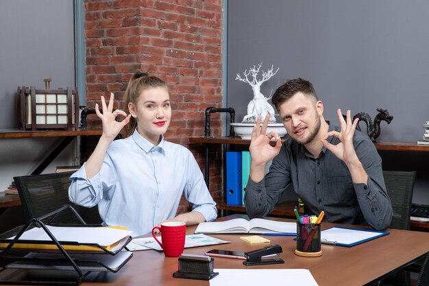 Bovenaanzicht van glimlachende en gemotiveerde geschoolde arbeiders die een brilgebaar maken in een kantooromgeving