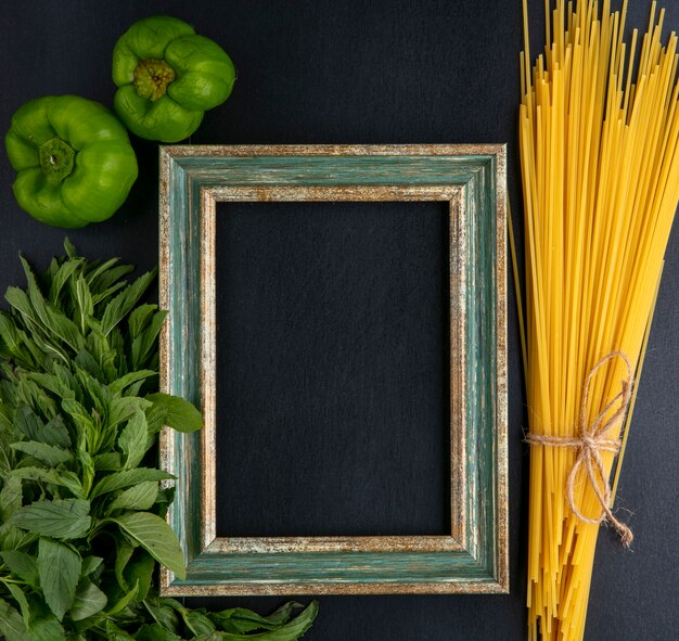 Bovenaanzicht van groenachtig-gouden frame met rauwe spaghettimunt en paprika op een zwarte ondergrond