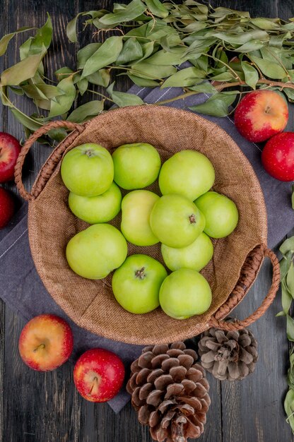 Bovenaanzicht van groene appels in mand met rode appels dennenappels en bladeren op doek en houten tafel