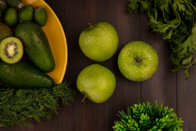 Bovenaanzicht van groene appels met vers fruit zoals avocado's feijoas en kiwi's op een gele plaat op een houten muur