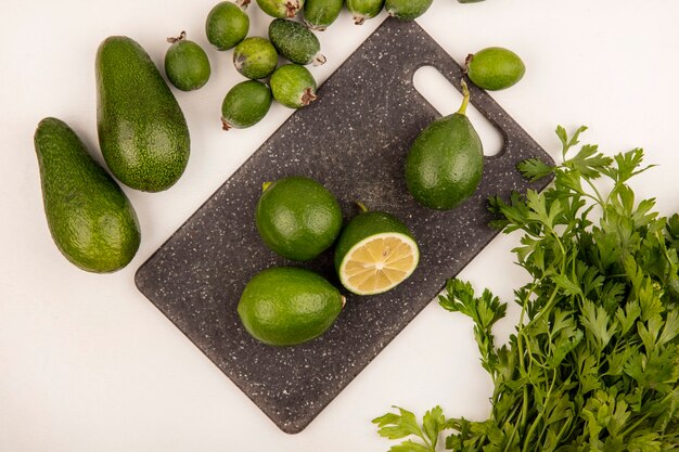 Bovenaanzicht van groene citrusvruchten limoenen op een keukenbord met feijoas avocado's en peterselie geïsoleerd op een witte muur