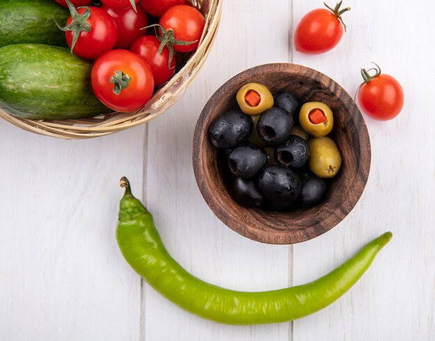 Bovenaanzicht van groene en zwarte olijven in kom en tomaten komkommers in mand en peper op houten oppervlak