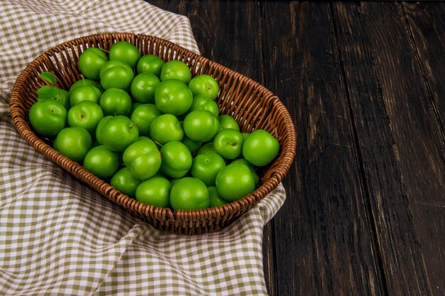 Bovenaanzicht van groene zure pruimen in een rieten mand op geruite stof op donkere houten tafel met kopie ruimte