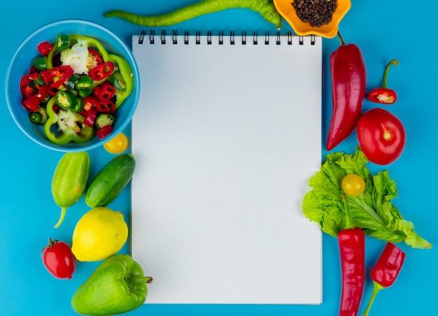 Bovenaanzicht van groenten als komkommer peper tomatensla met zwarte peper zaden en gesneden paprika's met notitieblok op blauw oppervlak met kopie ruimte