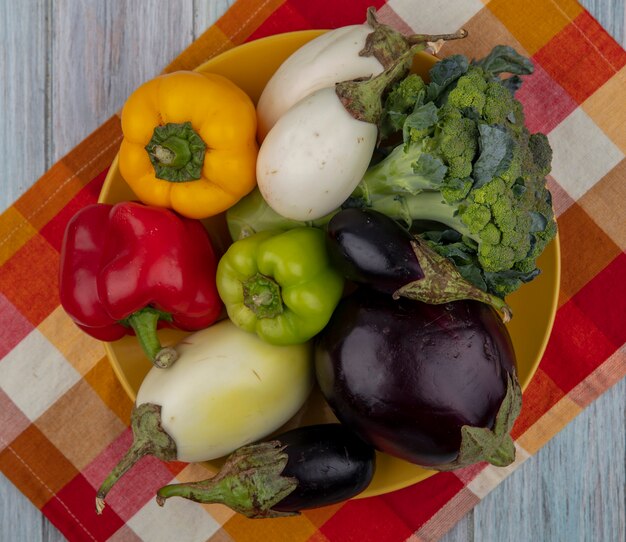 Gratis foto bovenaanzicht van groenten als peper, broccoli en aubergine in plaat op geruite doek op houten achtergrond