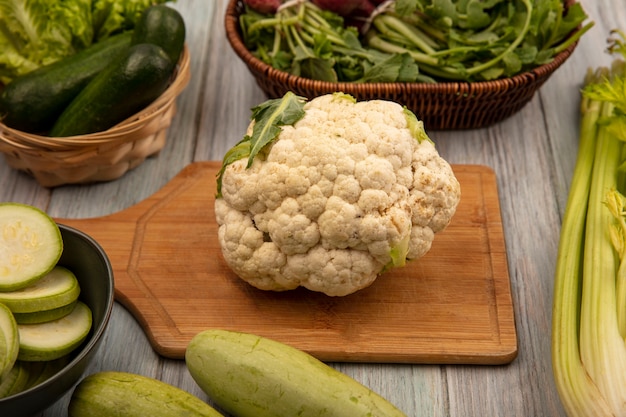 Bovenaanzicht van grote witte en ronde plantaardige bloemkool op een houten keukenbord met gehakte courgettes op een kom met komkommers en sla op een emmer met selderij en courgettes