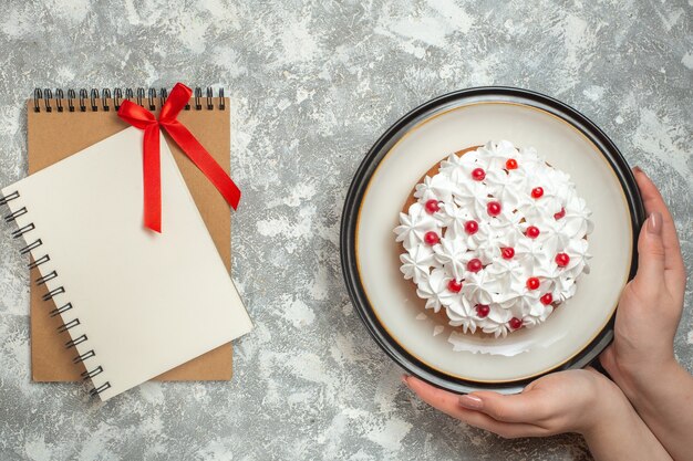 Bovenaanzicht van hand met een bord met heerlijke romige cake versierd met fruit naast notitieboekjes op ijsachtergrond